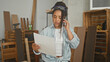 © Krakenimages.com - Woman carpenter holding smartphone to ear and reading a sheet of plans in studio workshop amid wooden planks and crates; concentration planning.