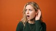 © haris - A surprised young blonde woman in a green sweater intently listens with her hand cupped to her ear against an orange background.