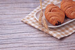 © Nurma - Golden croissants rest on a white plate. They sit atop a checkered napkin. Two wheat stalks lean beside the pastry. Wooden surface adds warm, rustic charm. Soft lighting highlights flaky