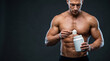 © mamo studios - Muscular shirtless bodybuilder preparing protein powder shake holding blank white supplement tub and scoop on dark background for sports nutrition fitness muscle growth and healthy lifestyle concept