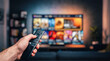 © Celt Studio - Person holding black remote control pointed directly towards large television display screen showing video streaming application inside dim modern living room