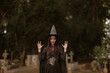 © Witoon - Gothic witch woman wearing a pointed hat and black costume, posing in a cemetery with a mysterious and eerie Halloween atmosphere.