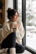 © Ksu_Sha - Smiling woman enjoying a warm drink by snowy window during a cozy and relaxing winter morning