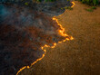 © Amazing Aerial - Aerial view of a wildfire spreading across dry grasslands with bright orange flames consuming vegetation and leaving behind charred black earth in Tomia, South East Sulawesi, Indonesia.
