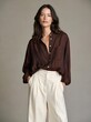 © Elena - Young woman standing in front of a plain grey background. she is wearing a brown blouse with a button-down collar and long sleeves. the blouse has a subtle pattern of small white dots on the front.