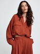 © Elena - Young woman with long dark hair, standing against a plain white background. she is wearing a long-sleeved, orange-colored top with a deep v-neckline and multiple pockets on the front.