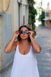 © dsbedarieva - A woman in a white dress adjusts her sunglasses while walking in Kerkira, Greece. Historic buildings and a stone street frame the narrow city alley behind her.