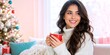 © Vasiliy - Smiling young woman holding a red mug in a cozy living room. Happy female in white sweater near a decorated Christmas tree. Winter holiday lifestyle concept