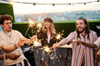 © pressmaster - Multiethnic group of happy young adults holding sparklers during rooftop party celebrating event. Perfect for friendship campaigns, social media marketing, and youth lifestyle branding