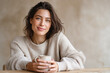 © Irina - Smiling brunette woman in a cozy beige sweater holding a warm drink at a wooden table in a softly lit neutral interior.