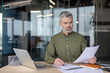 © Liubomir - Businessman working in office, mature professional man reviewing documents and writing on paper at desk with laptop for financial analysis tasks. Focused executive managing business strategy.