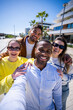 © CarlosBarquero - Vertical. Four cheerful friends gather closely for a selfie in a bright residential promenade, enjoying friendship and a sunny day together outdoors.