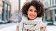 © Iryna - Smiling young woman with curly hair wearing a warm knitted scarf and coat outdoors on a snowy winter day in the city