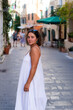 © dsbedarieva - A young woman wearing a white dress walks along an outdoor street in Kerkira, Greece. Traditional multi-story buildings line the background of the paved lane.
