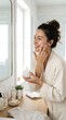 © Wellington - A woman in a bathrobe applies cream to her face in a bright, airy bathroom, showcasing a moment of self-care and beauty routine.