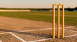 © bayu - Traditional Wooden Cricket Stumps and Bails on Pitch Crease at Sunset in an Outdoor Sport Stadium
