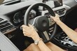 © Aleksei Isachenko/imageBROKER - A person grips the steering wheel of a modern vehicle, focusing on the dashboard display while driving through a vibrant city. Sunshine fills the interior, highlighting sleek design features