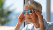 © Nataliya - Older woman wearing sunglasses indoors near a sunny window, gently rubbing her temples, light sensitivity and headache concept, migraine awareness, senior healthcare, realistic