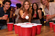 © Sunny - Low table-level view of red cups arranged for beer pong as a young woman takes her shot and friends react around the game table.