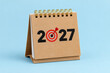 © Wasif - Desktop calendar showing year twenty twenty seven with a red dart hitting the bullseye center symbolizing future business goals and strategic planning success isolated on a bright blue studio backgrou
