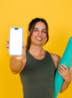 © Daniel - Smiling young woman holding a smartphone with a blank white screen for mockup while carrying a rolled yoga mat, presenting fitness app or online workout content