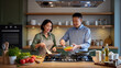 ©  VladaToday - Smiling couple cooking together in a bright modern kitchen, woman stirring vegetables in a skillet while man adds chopped ingredients from a glass bowl at the stove