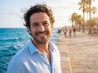 © Pixel Stock House - Cheerful young man with curly hair smiling on coastal promenade at sunset, portrait of attractive male tourist enjoying summer travel vacation along seaside boardwalk with palm trees blur
