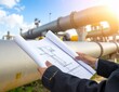 © AzamGold - Engineer Reviewing Construction Plans with Industrial Pipelines in the Background Under Clear Sky