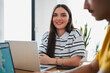 © Studio Marmellata - A young smiling woman sits at a wooden desk with a laptop in a bright modern office. She looks directly at the camera while collaborating with a colleague.