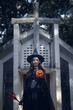 © ThawKyar - Halloween theme: A young woman with ghostly makeup and hair, dressed as a witch, holding a magic wand and a pumpkin amidst a graveyard; Halloween festival concept.