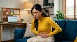 © BrilliantPixels - Young businesswoman experiencing severe menstrual cramps while sitting on a blue sofa.