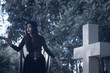 © ThawKyar - Halloween theme: A young woman with ghostly makeup and hair, dressed as a witch, holding a magic wand and a pumpkin amidst a graveyard; Halloween festival concept.