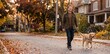 © ProNoJoy - A smiling man in a brown jacket walks his golden retriever dog along a suburban street covered in fallen autumn leaves, with residential houses and trees in the background, during golden hour