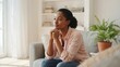 © Hosnayara - Thoughtful young black woman sitting on sofa, looking away and contemplating in a bright living room