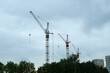 © syntheticmessiah - Construction background. A row of tall tower cranes silhouetted against a grey cloudy sky. Green trees in the foreground and apartment buildings on the right