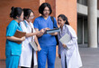 © Prot - Asian healthcare students in white coats and scrubs discuss coursework together outside a university building, using tablets and notebooks in a collaborative campus learning setting.