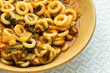 © Fotema - Close-up of an authentic Italian Anelli Siciliani con Bietole e Pomodori dish. A brown ceramic plate with ring-shaped pasta on a bright background featuring a beige geometric pattern.