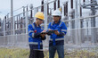 © Father_Studio - Two electrical technicians stand beside a fenced power substation as one communicates by handheld radio while the other holds inspection documents during outdoor maintenance and safety work.
