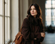 © Anna - Elegant brunette woman in layered brown knitwear stands beside a large window with a leather shoulder bag, gazing thoughtfully into the distance.