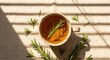 © Sara - A soothing cup of herbal rosemary tea in a ceramic mug on a beige linen tablecloth, illuminated by warm morning sunlight through window blinds creating beautiful shadows for a wellness concept.