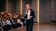 © Azorahaiá - Confident Woman Speaker Addresses Audience in Modern Auditorium Holding Microphone