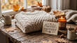 © Arra Vais - Sweater Weather card and folded knitted sweater on rustic wooden table. Autumn composition with glowing candle and hot drink in mugs near window