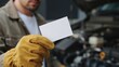 © Ruby - Mechanic wearing yellow leather work gloves holding a blank white business card in front of an open car engine hood in a garage workshop