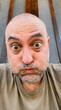 © Grey Zone - A close-up selfie of a middle-aged bald man with a beard making a funny face with wide eyes and pursed lips against a textured glass background.