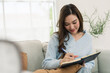 © Ekkasit A Siam - Young Asian woman records daily note in diary book on sofa, relaxed home lifestyle, thoughtful journaling session, weekend planning habit, bright apartment comfort, mindful quiet time indoors.