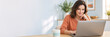 © Milan - Smiling young woman working on a laptop at home. Female freelancer or student sitting at a desk in a modern home office. Copy space for text