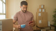 © Krakenimages.com - Man holding smartphone and coffee mug while checking orders among stacked boxes in a packing building; calm focus.