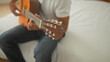 © Krakenimages.com - Man strumming acoustic guitar on bed in studio, hands fingering chords and focused expression while seated on white bedding and orange cushions; concentration.