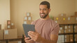 © Krakenimages.com - Man holding tablet, smiles and waves amid stacked parcels in a small business warehouse; entrepreneurial growth happiness.