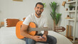 © Krakenimages.com - Man with acoustic guitar and smartphone, strumming and smiling while seated on bed in a cozy home studio with plants and shelves; relaxed practice.